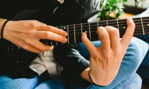 Dłonie grające na czarnej gitarze elektrycznej, z widocznymi progami i strunami.