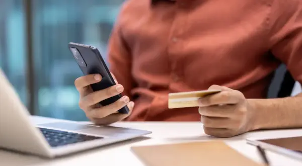 Osoba trzymająca telefon i kartę kredytową przy laptopie.