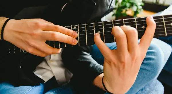 Dłonie grające na czarnej gitarze elektrycznej, z widocznymi progami i strunami.